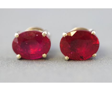 A white metal pair of single stone stud earrings. Each set with an oval faceted cut composite (glass filled) ruby. Post and s