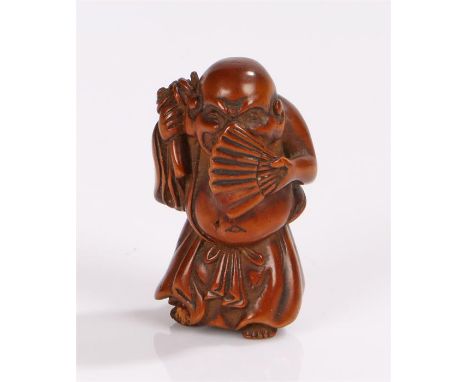 Japanese Meiji period boxwood netsuke depicting a standing figure of Hotei his face partial hidden by a fan, with his bag to 