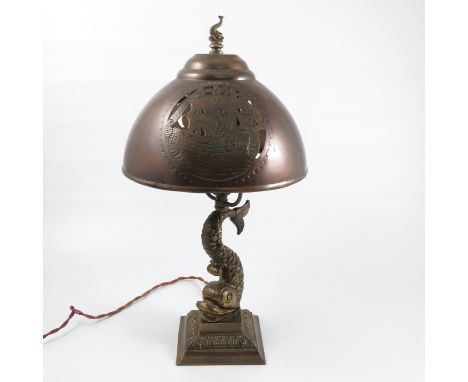 A brass table lamp, the column formed as a fish to a stepped square base with leaf decoration, the metal shade pierced and en