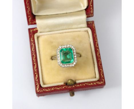 An early 20th century untested emerald and diamond cluster ring, the central emerald cut emerald within a white metal milgrai