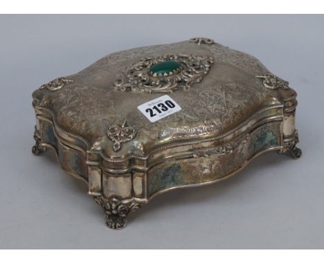 A shaped rectangular hinge lidded trinket box, the hinged lid with a cabochon green gem set centre, within a scrolling surrou