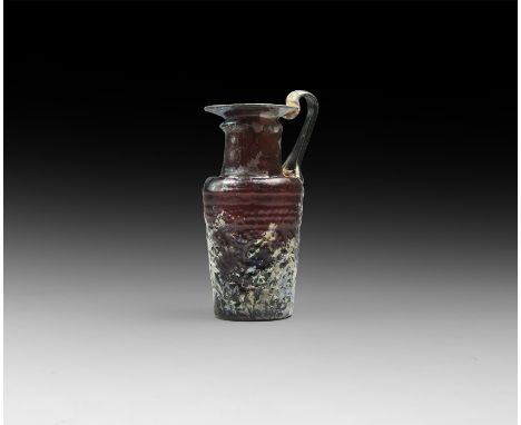 5th-7th century AD. An aubergine-coloured glass jug comprising a tapering body and flat base, carinated shoulder, broad tubul