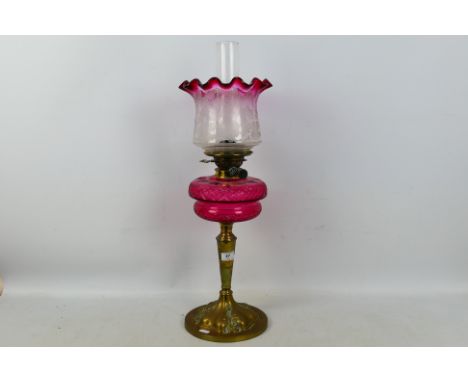 A brass oil lamp with cranberry glass font (font approximately 19 cm diameter) and cranberry tinted shade, height 66 cm.Condi