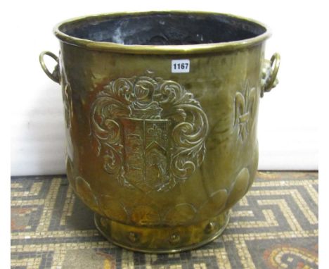 An embossed brass coal/log bin of cylindrical form with lions mask and ring handles, armorial shield and further decoration a