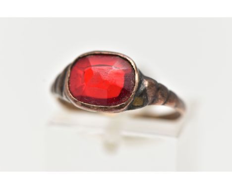 A YELLOW METAL GEM SET RING, set with an oval cut red stone assessed as paste, collet set in a yellow metal, textured shoulde