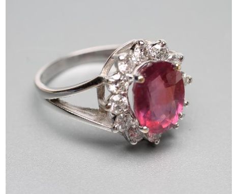 A RUBY AND DIAMOND CLUSTER RING, the oval facet cut ruby claw set to a border of twelve brilliant cut diamonds to open should