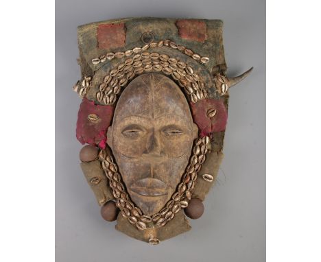 A West African tribal Dan hardwood mask featuring fabric and leather head cover heavily decorated with cowrie shells, feather