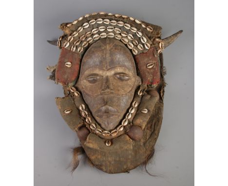 A West African tribal Dan hardwood mask featuring fabric and leather head cover heavily decorated with cowrie shells, feather