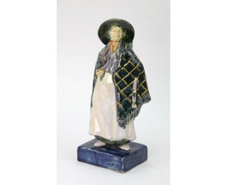 A ceramic figure of a woman wearing a cape and a wide brimmed hat on a rectangular blue plinth base, initialled 'R W' and dat