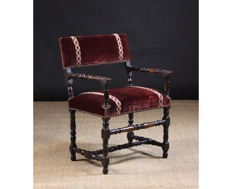 A 17th Century French Walnut Armchair, The padded rectangular back panel and seat covered in crimson velvet banded with braid