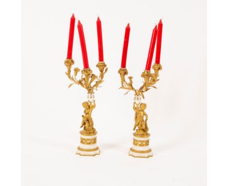 A pair of ormolu three-branch, three-light table lights with putto supports on white and gilded marble bases, 36cm high