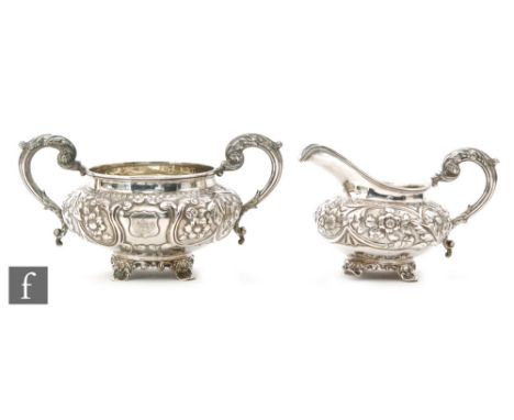 A hallmarked silver cream jug and sugar basin each with embossed foliate decoration around a central crest, each terminating 