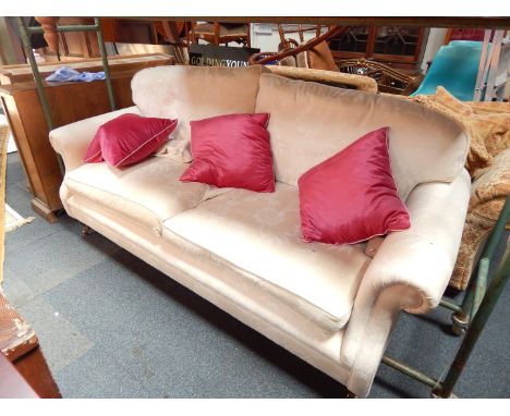 A two seater sofa with scroll arms and loose cushions, upholstered in a cream fabric, raised on turned legs, brass capped on 