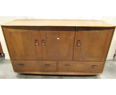 An Ercol medium to light elm sideboard enclosed by three doors over two drawers with oval dished handles, raised on shepherds