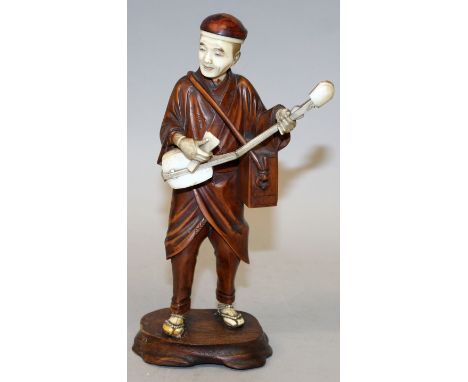 A JAPANESE MEIJI PERIOD WOOD & IVORY OKIMONO OF A STREET MUSICIAN, standing and playing a samisen, 8in high.