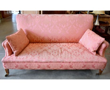 An Edwardian Howard style sofa with straight back and shaped arms, mahogany legs and brass casters, and pink damask upholster