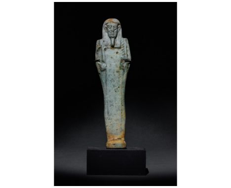 Ca. 664-332 BC. Late Period. A pale blue-glazed faience shabti on a modern stand. The head wears a striated lappet wig and a 