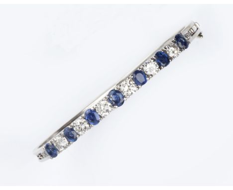 A SAPPHIRE AND DIAMOND BANGLE, of hinged bifurcated form, set to the top with a line of alternate oval mixed-cut sapphires an