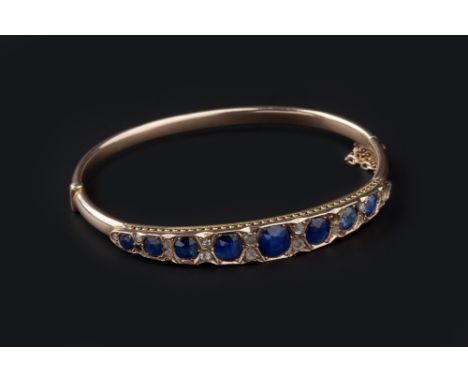 A SAPPHIRE AND DIAMOND SET BANGLE, of hinged oval form, applied with a tapered panel of graduated cushion-shaped sapphires, s