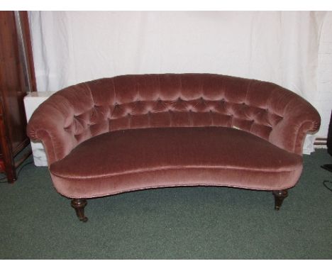 Victorian curved sofa with a dusty pink button back upholstery on tapering mahogany legs, the brass castors marked Shoolbred 