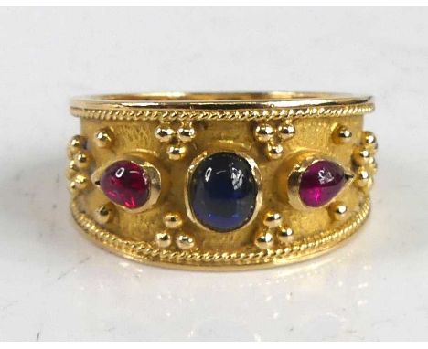 A yellow metal Etruscan style dress ring, having a centre oval synthetic sapphire and two synthetic pear shaped ruby cabochon