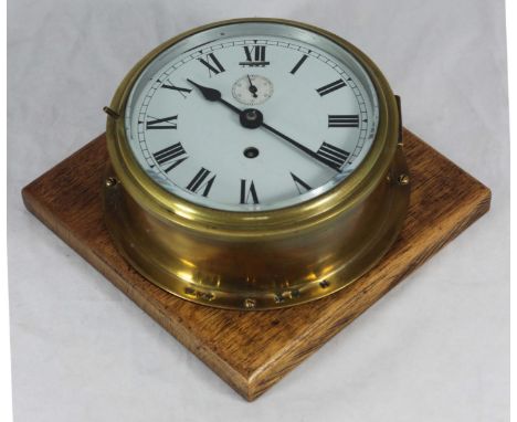 A brass bulkhead clock, the white enamel dial with Roman numerals denoting hours and subsidiary seconds dial, mounted on oak 