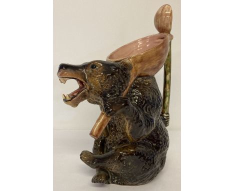 A large vintage ceramic majolica glaze honey bear jug with large spoon shaped handle. Dark brown coloured bear holding a pink