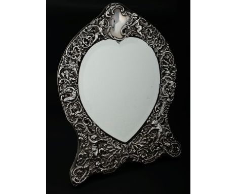 A Victorian silver table mirror by William Comyns, the cartouche-shaped frame profusely decorated with putti amidst scrolling