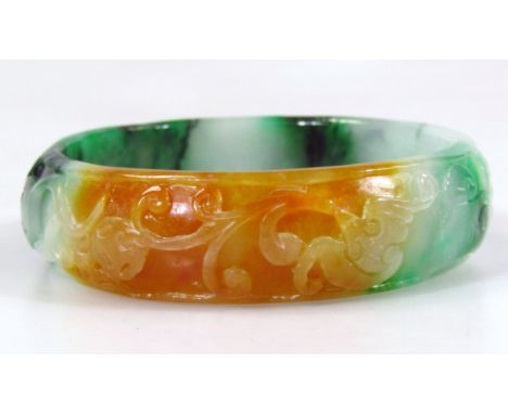 A Burmese style tri-colour jade bangle, raised with a repeat scroll and flowerhead pattern, predominantly in green, orange an
