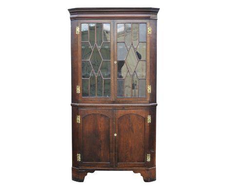 A George III oak standing corner cupboard, in two sections, the upper glazed panel doors above two arched panel doors, on bra