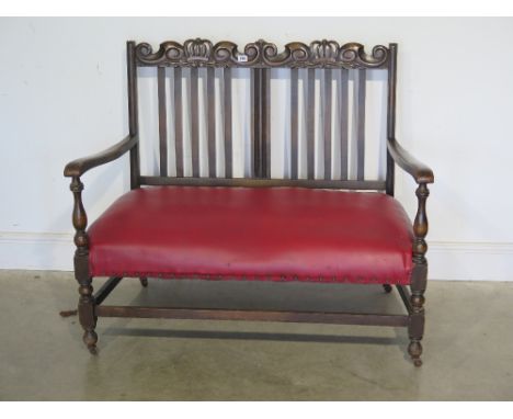 A carved oak two seater bench, crowns and scrolls to back above red leatherette seat, raised on turned supports and joined by