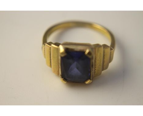 A yellow metal (tests as 14 carat) dress ring with stepped shoulders, set to centre with an emerald cut blue paste stone. Gro