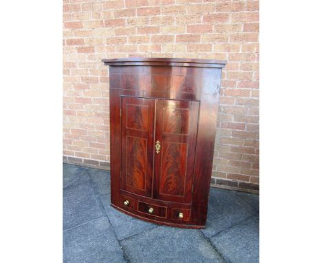 A GEORGIAN MAHOGANY BOW FRONT HANGING CORNER CUPBOARD the pair of panelled doors opening to reveal three shelves above three 