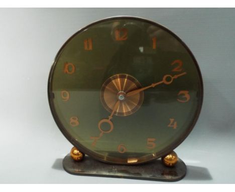 An Art Deco styled mantel clock, circular green glass dial mounted on an oval base, flanked by spherical brass decoration, 8-