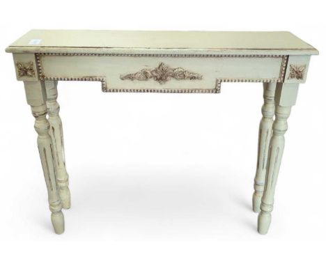 A narrow wooden console table, painted white with fading gold decorations. At the table's centre hangs a plate with leaf-like