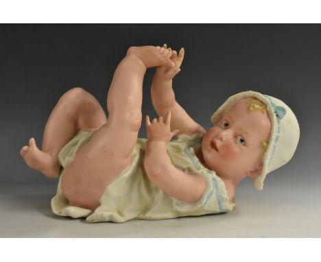 A large Gebruder Heubach bisque piano baby, on her back, playing with her feet, wearing a bonnet and cotton dress, 32cm long,