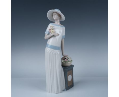 This elegant, tall figurine depicts a lady gracefully leaning on a basket of delicate flowers, gazing thoughtfully into the d