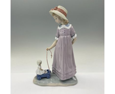Glazed porcelain figure of a young lady in a hat standing looking down at her doll in a small wagon. Lladro backstamp. This i