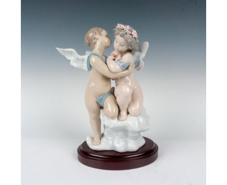 Glossy finish; two cherubs gracefully hug while sitting upon a cloud. This beautiful piece is displayed on a wooden base. Lla