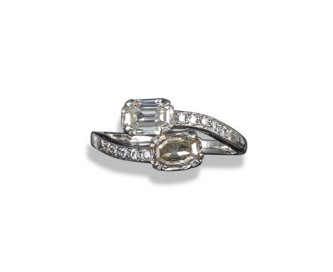 A two-stone diamond crossover ring, the two emerald-cut diamonds claw-set in platinum with further circular-cut diamonds to t