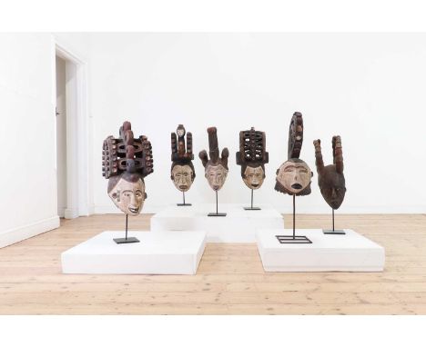 A set of six carved and painted wooden Igbo helmet masks, probably early 20th century, Nigerian, on wrought metal display sta