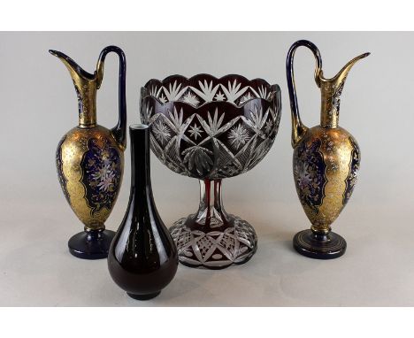 A ruby and clear overlaid glass bowl on stand, a ruby glass bottle vase, and a pair of ornate blue and gilt decorated jugs