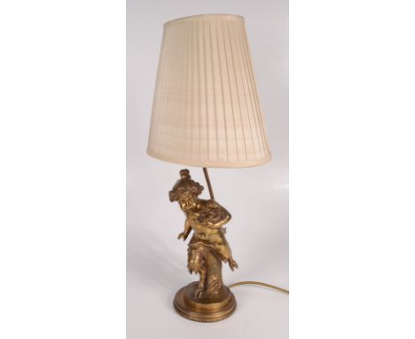 A gilt bronze table lamp, modelled as a child with cloven hooves, holding a bird's nest, with a cream linen pleated shade, fu