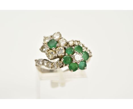 A DIAMOND AND EMERALD CROSSOVER RING, the white metal ring of crossover design, in the form of two flowers, one set with a ce