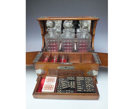 A LIGHT OAK THREE BOTTLE 'GAMING' TANTALUS,  with carved decoration, white metal mounts and carry handles, the two part hinge