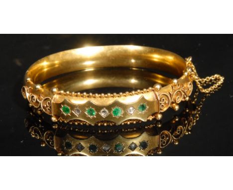 A Victorian style diamond and emerald set 9ct gold hinged bangle, the shaped rectangular panel set with seven alternating sto