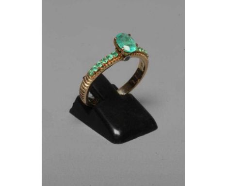 AN EMERALD DRESS RING, the central oval facet cut stone high claw set with a small diamond to each edge of the shank, with si