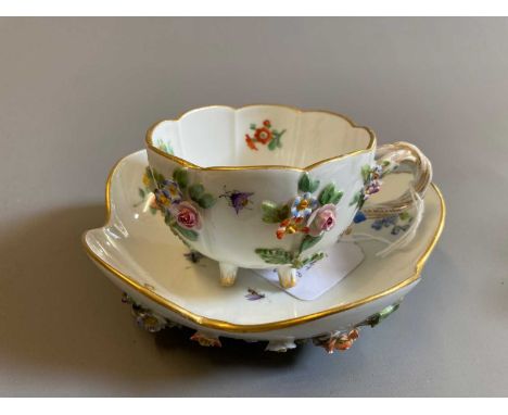 A MEISSEN PORCELAIN CABINET CUP AND SAUCER of lobed form raised upon four peg feet, encrusted and painted in polychrome ename