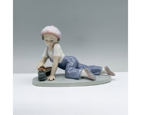 A young boy, clad in a lavender cap and denim overalls, happily engages with his toy train on the floor. Lladro backstamp. Th