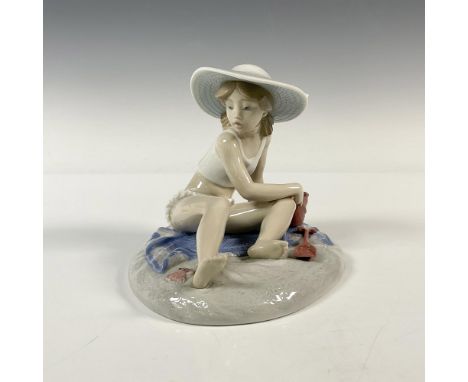 A young girl in a white beach outfit joyfully plays with her pail and shovel in the sand, sitting atop a blue towel. Lladro b